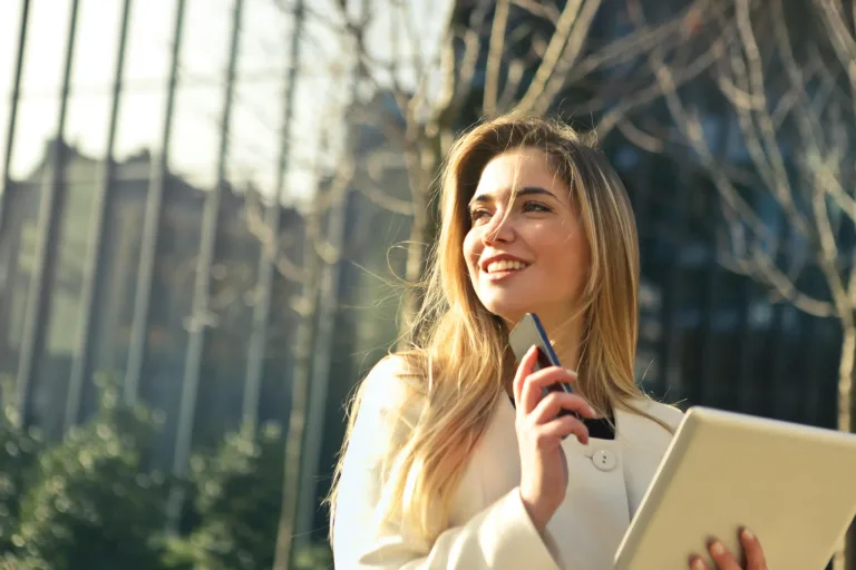 Woman in the sun with a phone and tablet in her hands. The woman looks to the side and smiles.